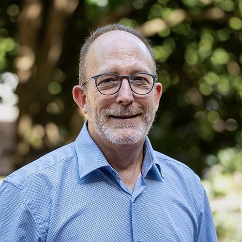 older man with short close-cropped hair, wearing glasses and light blue dress shirt while standing in front of a blurred tree in the background