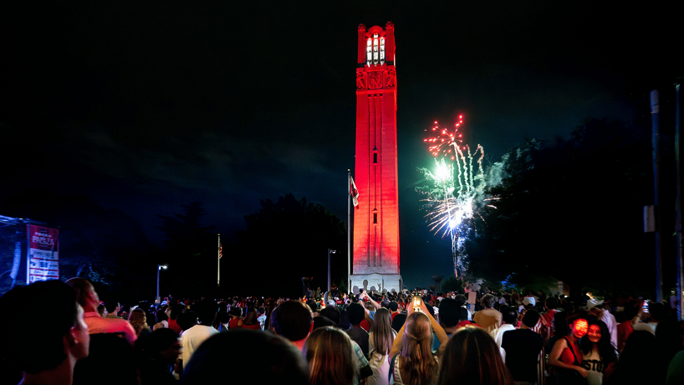 Image shows the NC State bell tower with fireworks in the background.