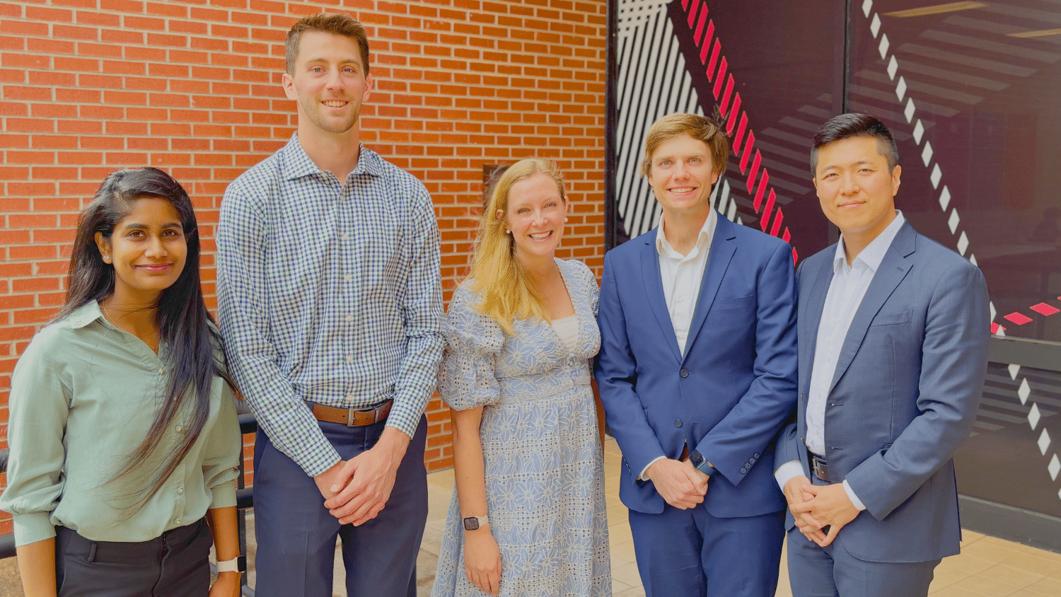 Image shows a group of professionally dressed men and women standing in front of a red brick wall