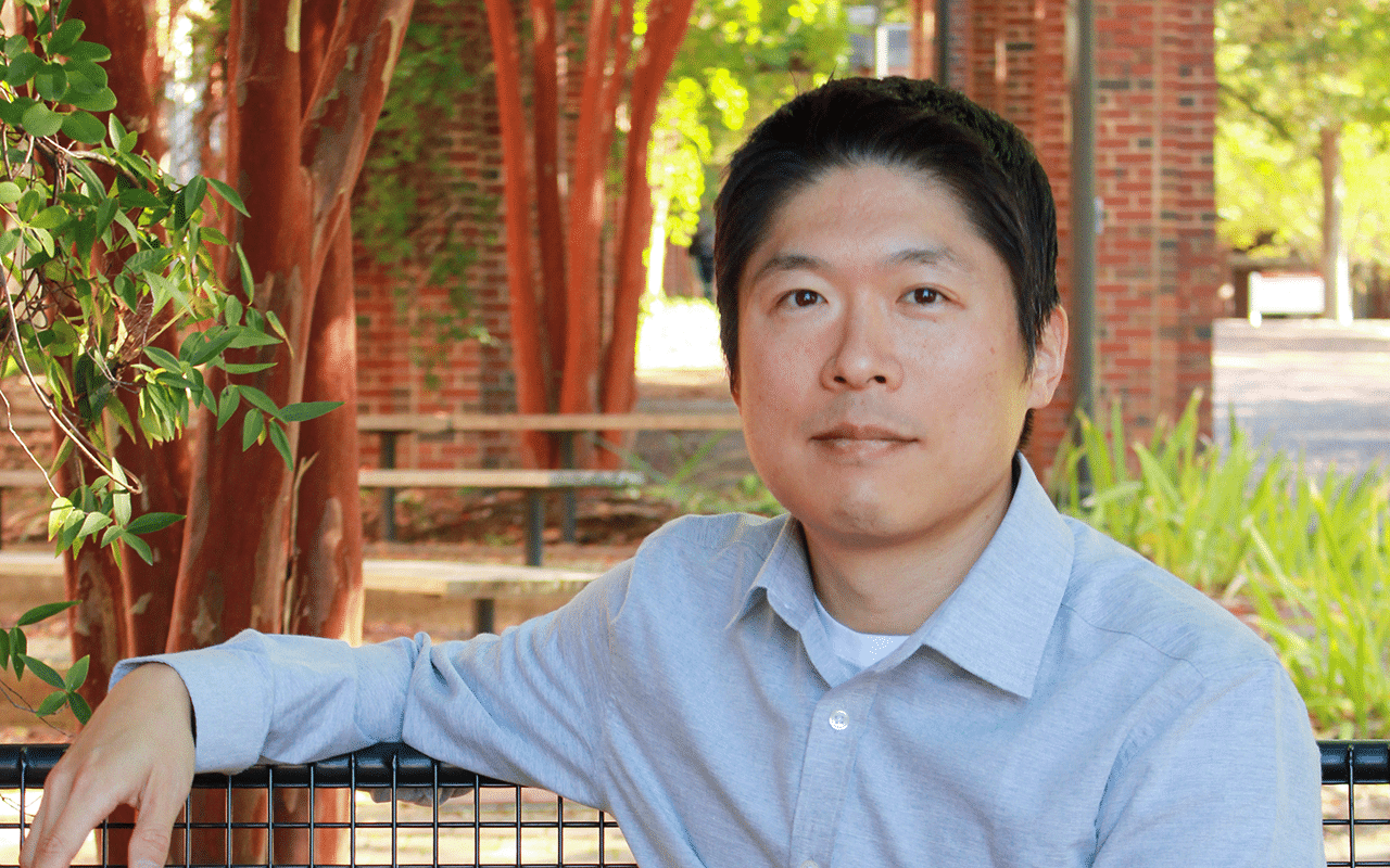 A man in a blue collared shirt with a college campus behind him.